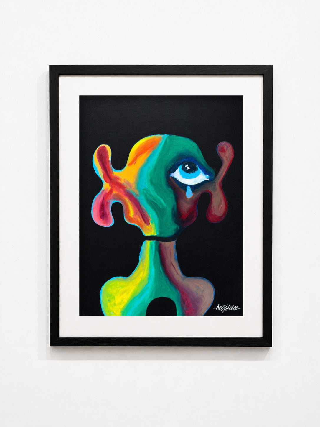 A framed abstract art print featuring a colorful, surreal figure with a single eye on a black background, presented in a black frame with a white mat against a white wall.